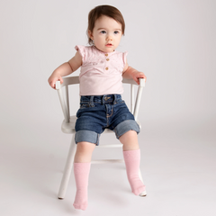 toddler with dark hair, light pink shirt, denim shorts and light pink socks sitting in a white chair